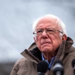 Elderly man in jacket speaking at outdoor event.