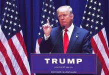 Man speaking at podium with American flags behind.