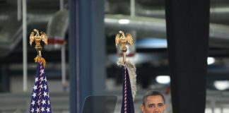 Man speaking at podium with flags behind him.