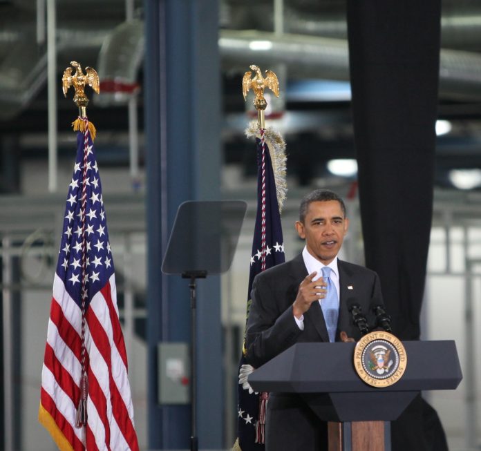 Man speaking at podium with flags behind him.