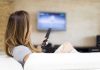 A woman sitting on a sofa, holding a remote control and watching television