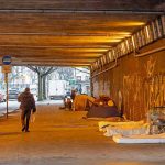 People walk under a bridge with homeless encampment.