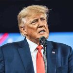 Trump Loyalist TURN – SLAMS President! Man in suit speaking at a microphone.