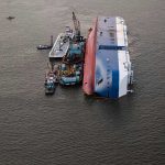Capsized ship with cranes and tugboats around it.