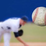 A baseball in mid-air with a pitcher in the background preparing to throw