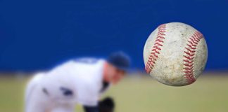 A baseball in mid-air with a pitcher in the background preparing to throw