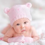 A cute baby wearing a pink knitted hat and mittens, lying on a soft surface
