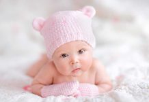 A cute baby wearing a pink knitted hat and mittens, lying on a soft surface