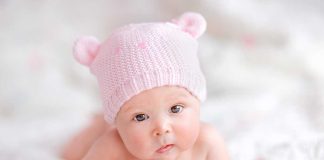 A cute baby wearing a pink knitted hat and mittens, lying on a soft surface