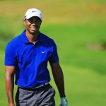 A smiling golfer wearing a blue shirt and white cap on a green golf course