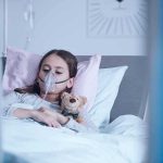 Child in hospital bed with oxygen mask holding teddy bear
