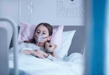 Child in hospital bed with oxygen mask holding teddy bear
