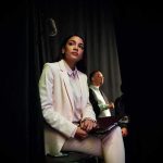 Trans Actor RAGES For Most Pathetic Reason Woman in a white suit sitting backstage with a folder