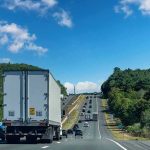 Semi-truck on multilane highway with cars and green hills under blue sky