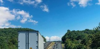 Semi-truck on multilane highway with cars and green hills under blue sky