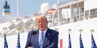 Donald Trump speaking at a podium with American flags and a ship in the background