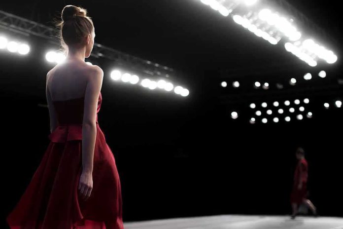 Model in a red dress walking on a runway under bright stage lights