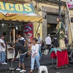 Street taco stand with people and a religious statue on a sidewalk