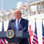 Controversial Trump Aide Appears Next To Him On Stage Donald Trump speaking at a podium with American flags and a ship in the background