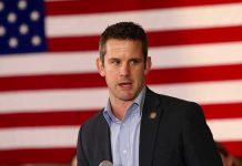 man speaking in front of a large American flag